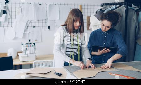 Young female tailors are busy working with tablet, communicating and outlining clothing pattern on textile on studio desk. Light workshop with sewing items in background. Stock Photo