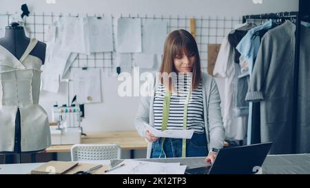 Young attractive woman is choosing sketches for new fashion show and using laptop in her modern tailor's shop. Creative thinking in clothing design industry concept. Stock Photo