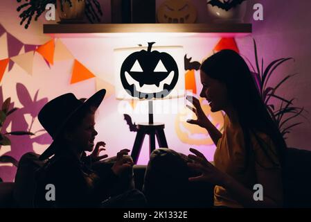 Silhouette of little daughter and young mother telling spooky stories sitting on cozy sofa in living room decorated for bats and pumpkins. Mom and kid Stock Photo