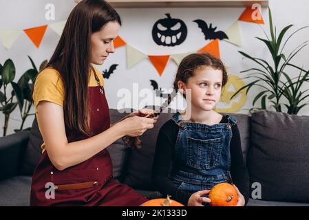 Caring young mother makes hairdo braids pigtail happy little kid daughter sitting on cozy couch in living room decorated spooky paper black pumpkins a Stock Photo