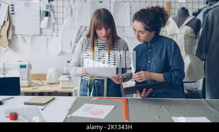 Creative clothing designers are looking at drawings and comparing them with images on tablet. Successful startup in fashion industry concept. Textiles, sewing machine and dummy are visible. Stock Photo