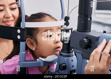 Checking little Korean girl's eyesight with binocular slit lamp in ophthalmology clinic, close-up. Vision correction in children Stock Photo