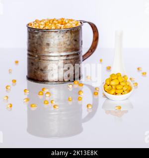 Cooked corn kernels on a white ceramic spoon with a metal container full of raw corn kernels in the background Stock Photo