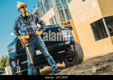 Caucasian Middle Aged Builder Proudly Standing in Front of Black Pickup Truck. Equipped with Hard Hat, Safety Headphones and a Hammer Hanging From His Stock Photo