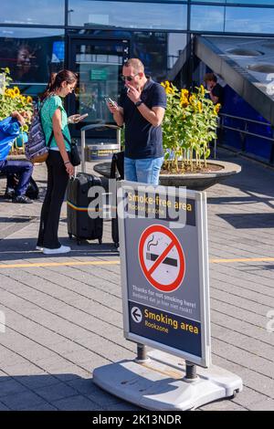 Two people smoking behind a sign stating this is a smoke-free area, and pointing to a smoking area, Schiphol Airport, Netherlands Stock Photo