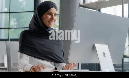 Young arab businesswoman in hijab working on computer smiling enjoying office work successful woman professional manager company employee develops Stock Photo