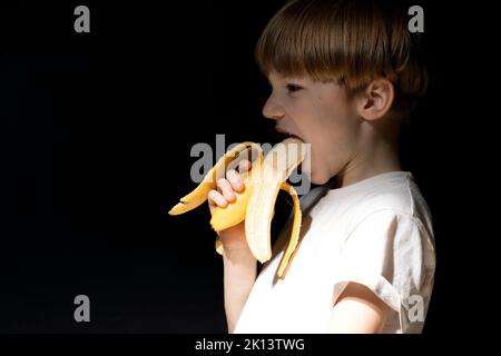 Funny portrait of boy with blond hair eating yellow banana on black background. Opened mouth. Studio shot Stock Photo