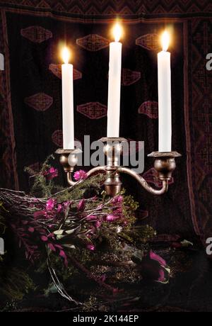 Concept image of three lighted candles in a silver candlestick holder, surrounded by a bouquet of dry and dying flowers, possibly denoting mourning Stock Photo