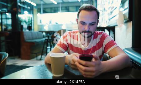 Young attractive man with a beard uses a smartphone in a cafe. Stock Photo