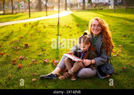 Redhead woman reads book with little boy sitting on grass in city park. Young mom teaches her first grader son and doing homework on sunny autumn day Stock Photo