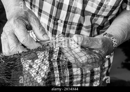 Close up of an old fisherman's textured hands mending a fishing net. Black and White Stock Photo