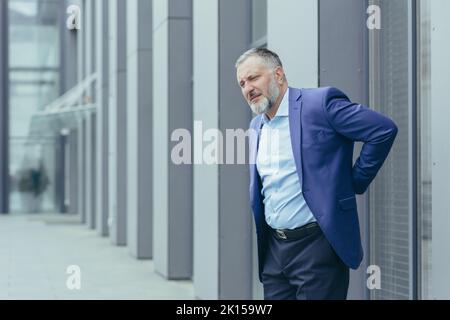 Senior male businessman suffering from back pain. Standing in a suit on the street near the office center, holding his hand behind his back, grimacing in pain, needs a break and medical help Stock Photo
