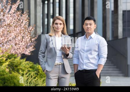 Portrait of two interracial businessmen. A young woman and a young Asian man are standing near the office in suit, Looking at the camera. A woman holds a tablet. A man holds his hands in his pockets. Stock Photo