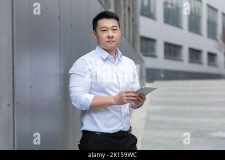 Portrait of a young male asian student standing on the street on the student campus. Holds a tablet in his hands, types, studies, prepares for exams. He looks seriously into the camera. Stock Photo