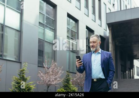 A senior man, a businessman in a suit, goes to a meeting on the street near a modern office center, holds a phone in his hand, looks at the phone, dials a number. He keeps his hand in his pocket. Stock Photo