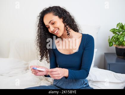 A young multi-ethnic woman sits on her bed holding pregnancy test and smiling Stock Photo