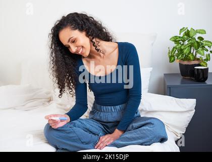A young multi-ethnic woman sits on her bed holding pregnancy test and smiling Stock Photo