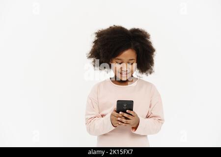 Black preteen girl with curly hair smiling and using cellphone isolated over white background Stock Photo