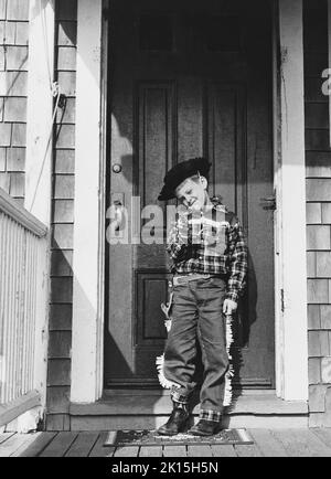 Six year old, Lewis E. Raymond, III, dressed as a cowboy brandishes his toy gun on the porch of his home. Many children participate in a form of role-playing known as make believe, wherein they adopt certain roles such as doctor and act out those roles in character. Sometimes make believe adopts an oppositional nature, resulting in games such as cops and robbers. Circa 1950-60s. Stock Photo
