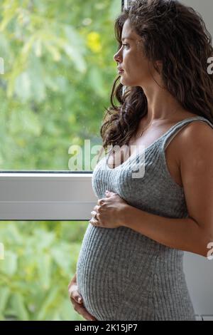 Beautiful pregnant woman in dress standing beside window and looking out with hands on belly Stock Photo