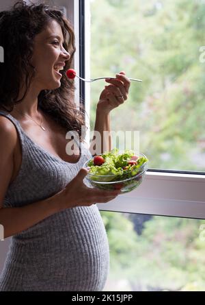 Pregnant woman standing beside window and eating fresh salad. Health care in maternity period Stock Photo