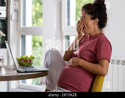 Pregnant woman yawning at table while working on laptop at home. Maternity leave and working from home concept Stock Photo