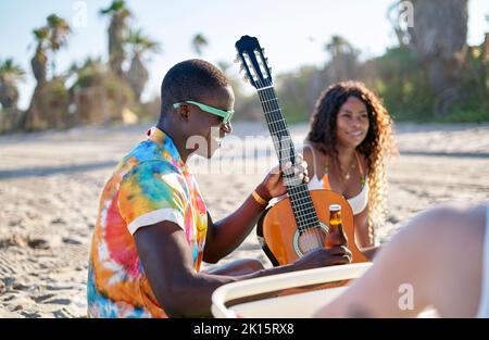 African American male musician in stylish colorful shirt and sunglasses drinking beer while sitting on sandy beach with acoustic guitar and talking to Stock Photo