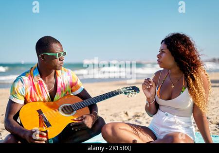 African American male musician in stylish colorful shirt and sunglasses drinking beer while sitting on sandy beach with acoustic guitar and talking to Stock Photo