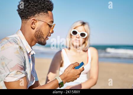 Black man sharing news in social media with girlfriend while spending time on sandy beach against sea and blue sky together Stock Photo