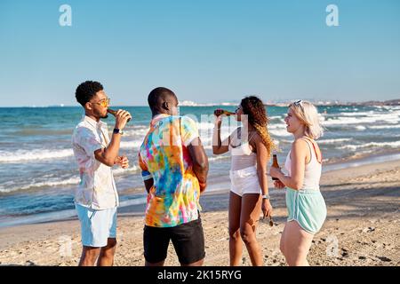 Positive young multiracial male and female best friends in summer clothes drinking beer and dancing on sandy beach on sunny day during vacation Stock Photo