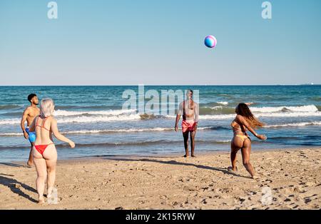 Black woman running and preparing to hit flying ball while playing with friends on sandy beach near sea Stock Photo