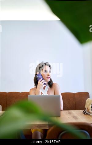 Female entrepreneur talking on mobile phone while using netbook and working remotely at table in cafe Stock Photo