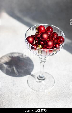 Holiday card concept with red Christmas balls inside champagne crystal glass on white concrete background with deep shadows Stock Photo