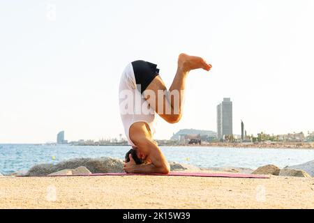 Full body unrecognizable barefoot male doing Salamba Sirsasana pose on beach near sea during yoga session in morning Stock Photo