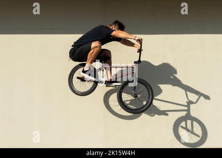 Full body side view male in casual clothes jumping on BMX bicycle against sunlit concrete wall on summer weekend day on street Stock Photo