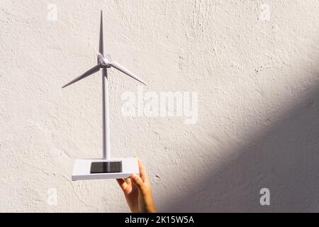 Crop unrecognizable person demonstrating small solar powered wind turbine toy against white concrete wall on sunny day Stock Photo