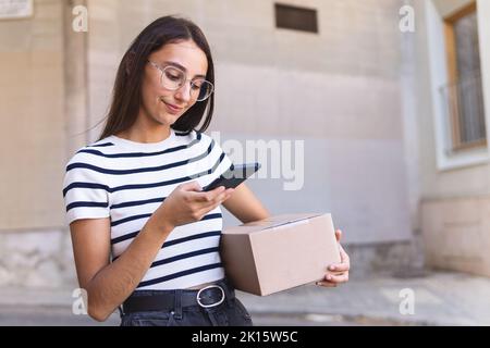 Crop cheerful young female courier with long dark hair in casual clothes and eyeglasses smiling and taking photo of cardboard box on smartphone while Stock Photo