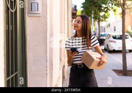 Positive young female courier in casual clothes and eyeglasses with cardboard box in hand using mobile phone while searching for address during delive Stock Photo
