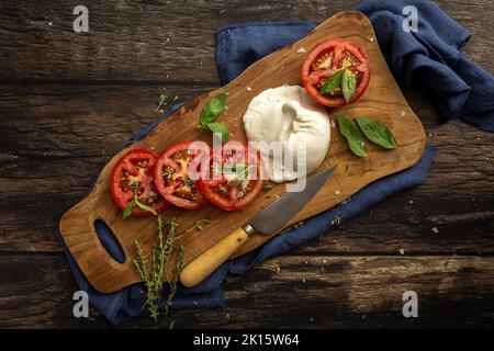 Top view of delicious mozzarella and slices of fresh tomato with green basil leaves and thyme served on wooden cutting board with knife on table Stock Photo