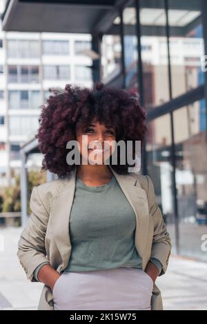 Positive black businesswoman in suit with curly hair holding hand in pocket while standing looking at camera on modern city street before work Stock Photo