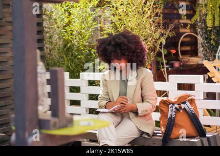 Thoughtful African American female entrepreneur in suit sitting on bench and writing in planner while working remotely on sunny day in street restaura Stock Photo