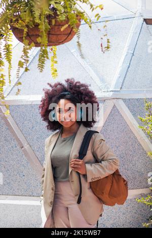 Optimistic young African American female manager in suit with backpack looking at camera with smile and listening to music while standing against geom Stock Photo