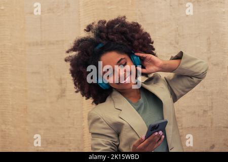 Merry black businesswoman in suit adjusting wireless headphones and browsing playlist on cellphone while standing against beige wall on street Stock Photo