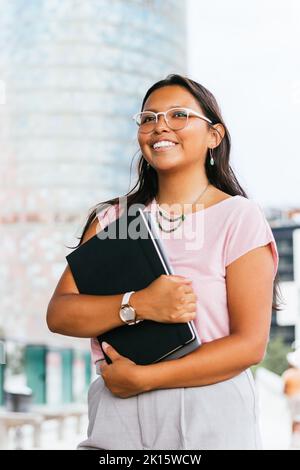 Cheerful young ethnic female student with long dark hair in casual clothes and eyeglasses smiling and looking away while embracing notebook and laptop Stock Photo