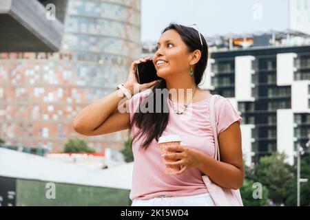 Cheerful young ethnic lady with long dark hair in casual clothes and eyeglasses smiling while talking phone and standing near modern building with cup Stock Photo