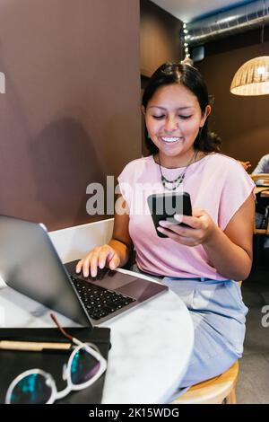 Happy young ethnic self employed lady with long dark hair in casual clothes smiling while messaging on mobile phone sitting at table with laptop durin Stock Photo