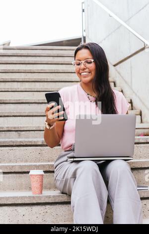 Positive ethnic female freelancer with dark hair in smart casual outfit and eyeglasses smiling and using smartphone while sitting on stairs with lapto Stock Photo