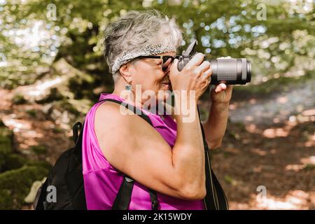 Side view of elderly female tourist with backpack shooting nature while spending sunny summer day in park Stock Photo
