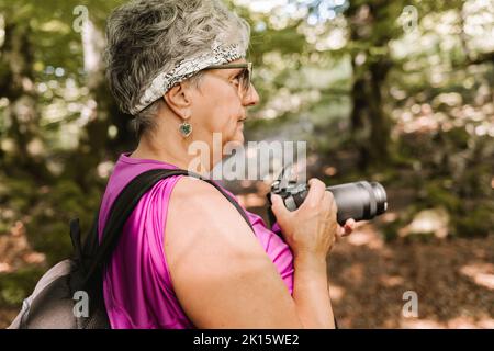 Side view of elderly female tourist with backpack shooting nature while spending sunny summer day in park Stock Photo