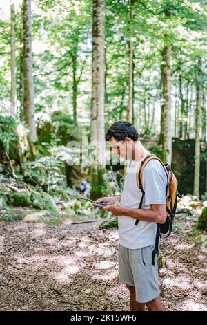 Man in casual clothes with backpack browsing GPS map on cellphone while exploring green forest on sunny summer day Stock Photo